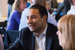 A person in business attire is smiling and engaged in conversation at a social gathering or meeting, with several people in the background.