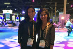 Two individuals are smiling at a professional event or conference. They are wearing name badges, and there are people and exhibition booths in the background.