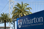 A sign for the Wharton School of the University of Pennsylvania is in the foreground, with palm trees and a bridge in the background.