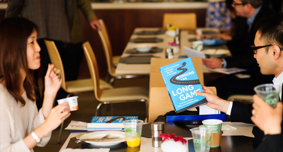 A group of people seated at a table in a restaurant or meeting room, with one person holding a book titled "Winning the Long Game."