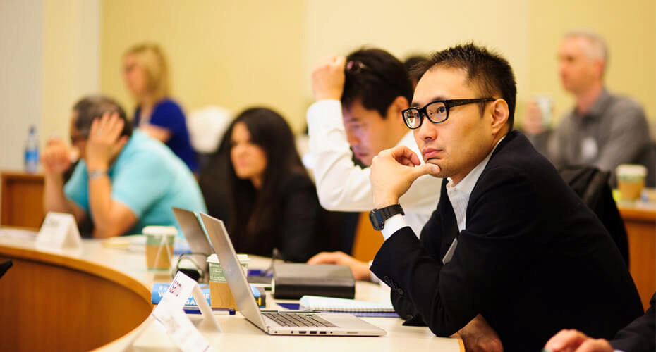A group of people in a classroom or seminar setting, seated at desks with laptops and notebooks, attentively listening to a presentation or lecture.