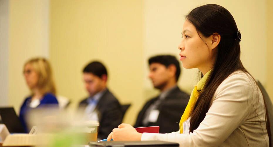 A small group of people sitting in a professional setting, focused on something ahead. One person in the foreground is wearing a yellow scarf.