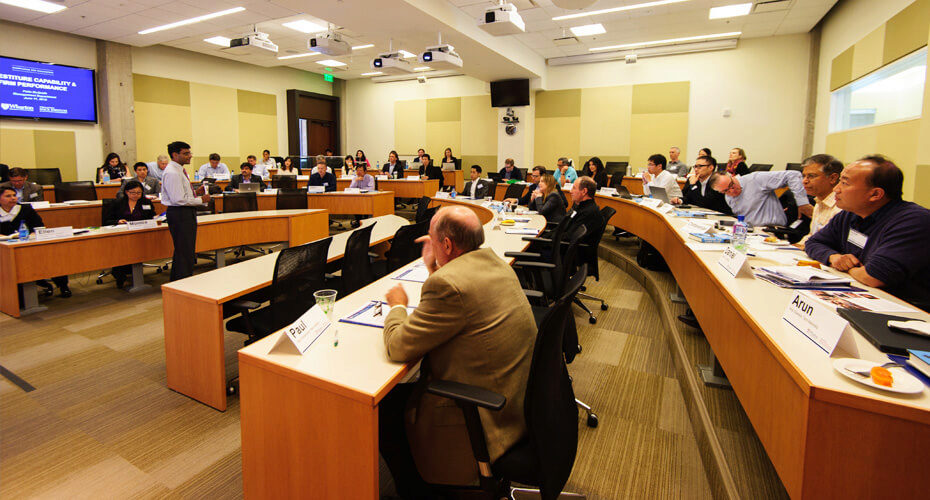 A classroom with several people engaged in a lecture or presentation. There are large desks arranged in a semi-circle, and a person is speaking at the front of the room.