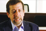 Headshot of a person with short hair and a beard, wearing a suit and shirt, speaking or in conversation in an indoor setting.
