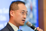 A headshot of a person speaking into a microphone, wearing glasses and a suit. The background is slightly blurred.