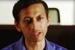 Headshot of a person wearing a blue shirt, speaking or in conversation, with a blurred background.