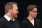 A couple of people in sweaters, sitting and looking to the side, against a dark background.