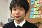 A person in a black sweater and white collared shirt stands in front of a wall covered with colorful sticky notes and pins.