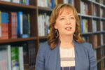 A headshot of a person with light brown hair wearing a blue jacket and earrings, standing in front of bookshelves filled with books.