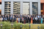 A large group of people dressed in formal attire posing for a group photo in front of a modern building with reflective windows.