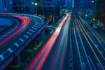 A night cityscape featuring light trails from moving vehicles on a highway and a curved overpass, surrounded by tall buildings.