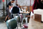 A cozy cafe setting with vintage chairs in the foreground, a small group seated around a table in the background, and a staff member serving.