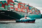 A large container ship loaded with colorful shipping containers on the water, accompanied by a small tugboat assisting in maneuvering.