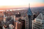 Aerial view of Philadelphia during sunset, featuring modern skyscrapers and historic buildings. The cityscape stretches towards the river in the distance.